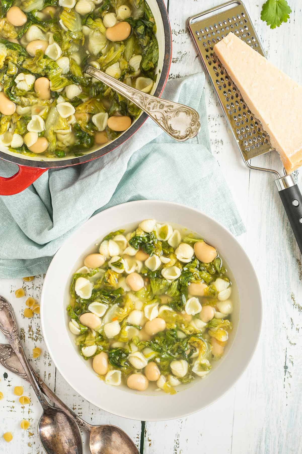 A white bowl of thin soup with white beans, chopped green escarole, and small shell pasta. Leftover soup is in a red enameled Dutch oven at the back.