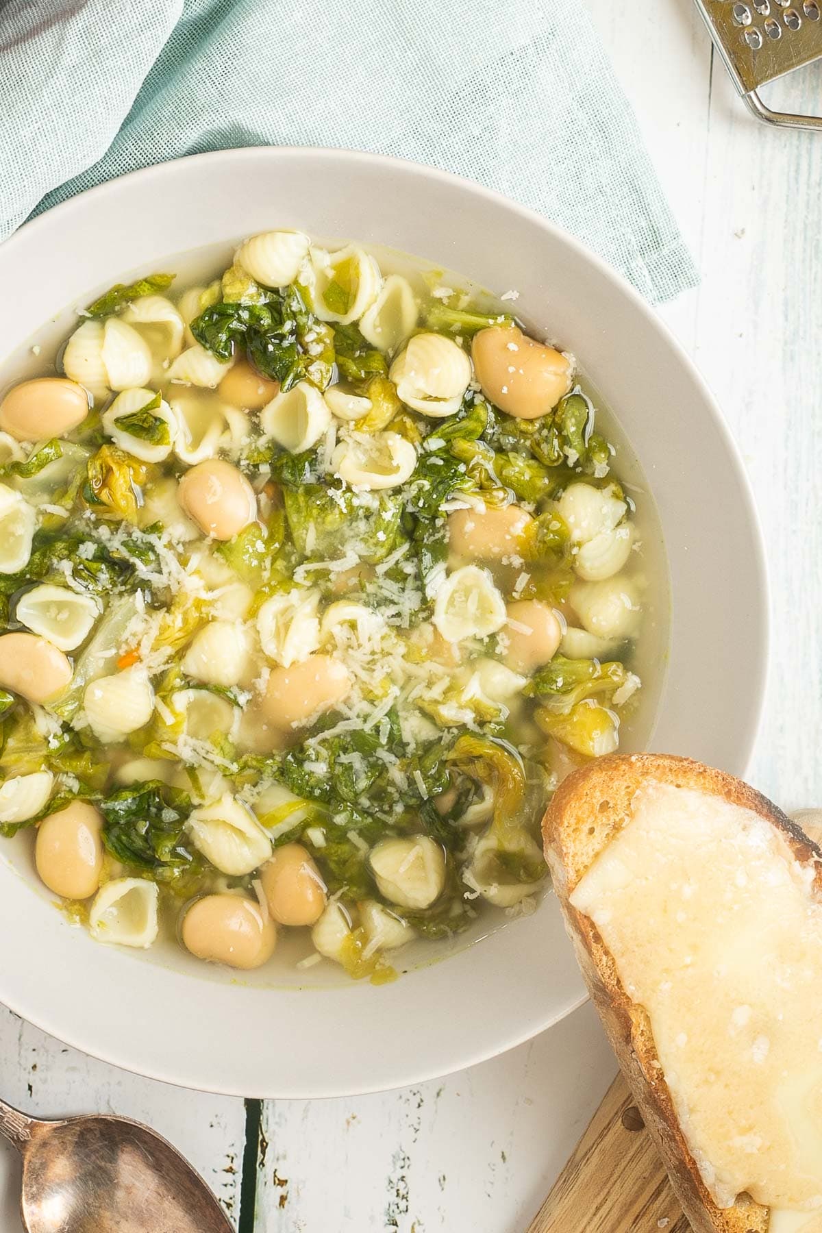 A white bowl of thin soup with white beans, chopped green escarole, small shell pasta, topped with grated cheese. A cheesy, toasted bread is placed on the side of the bowl.