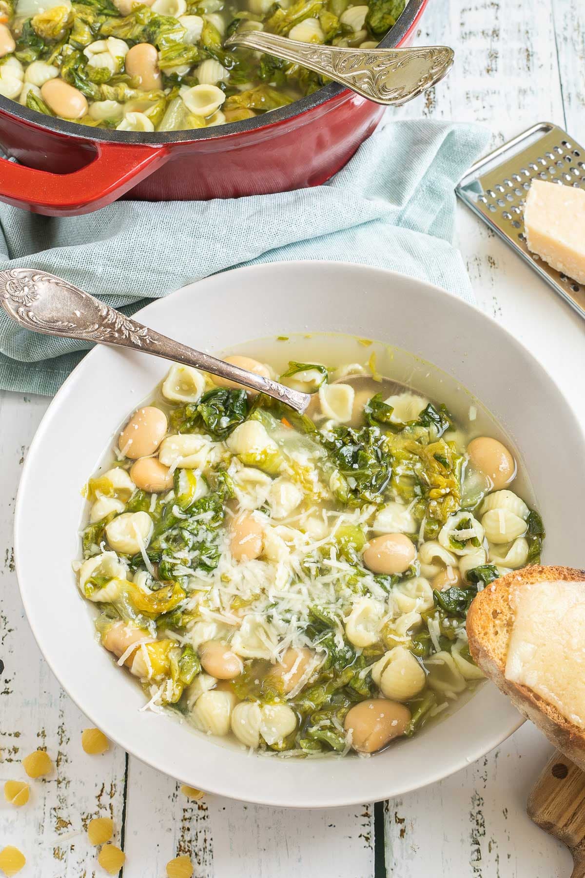 A white bowl of thin soup with white beans, chopped green escarole, small shell pasta, topped with grated cheese. Leftover soup is in a red enameled Dutch oven at the back. A cheesy, toasted bread is placed on the side of the bowl. 