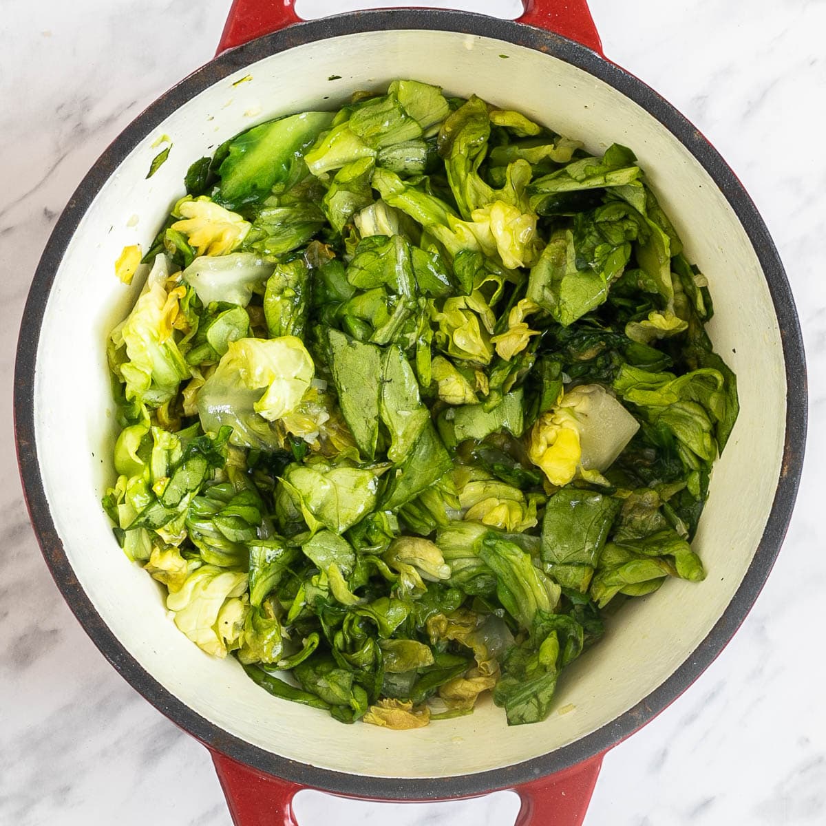 A red white enameled Dutch oven with chopped wilted green escarole.