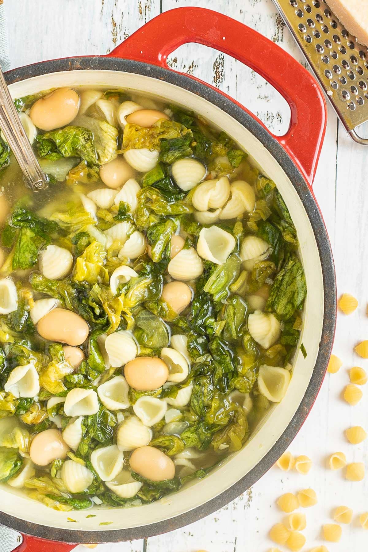 A red white enameled Dutch oven with a thin soup loaded with white beans, chopped green escarole, and small shell pasta.