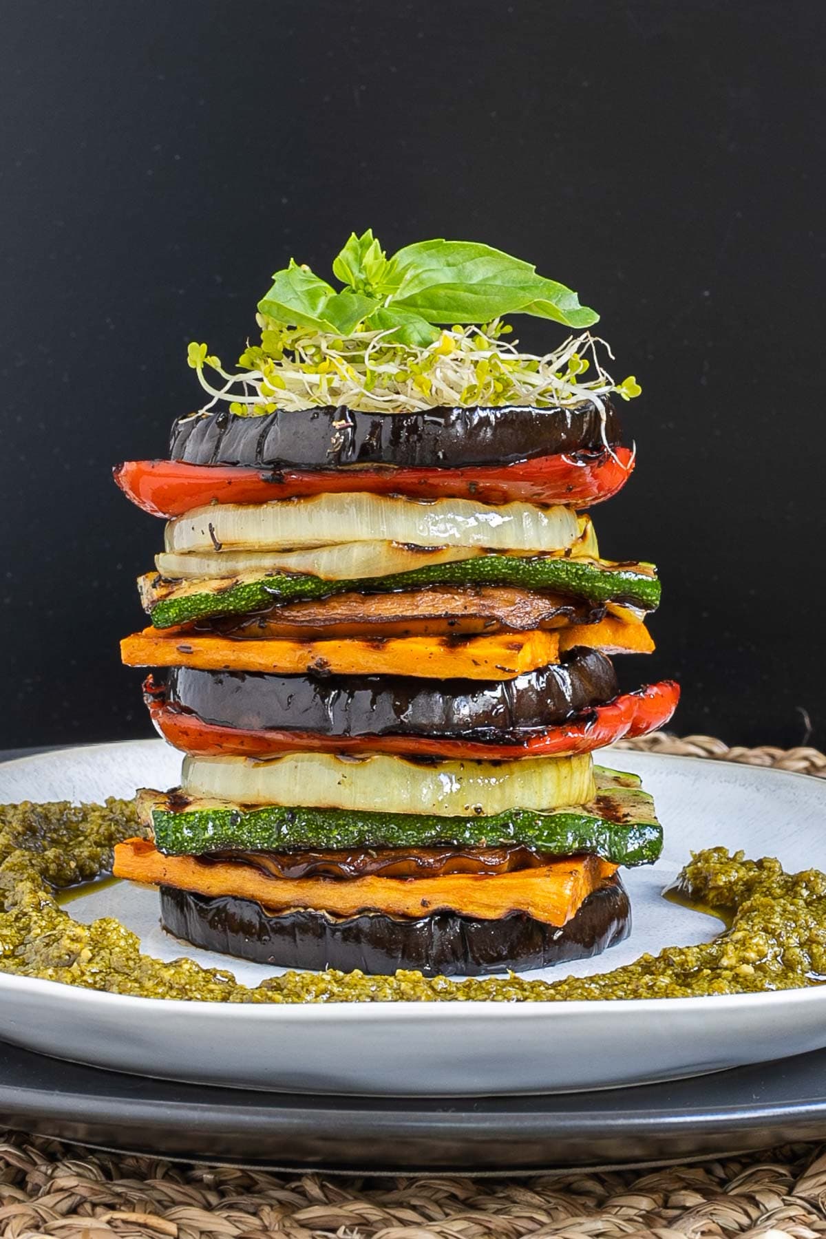 Different veggie slices are stack on top of each other to create a tower of veggies. It has eggplant, zucchini, sweet potato, onion, red bell pepper and mushroom slices. Green pesto is served around it. It is topped with sprouts and basil leaves.