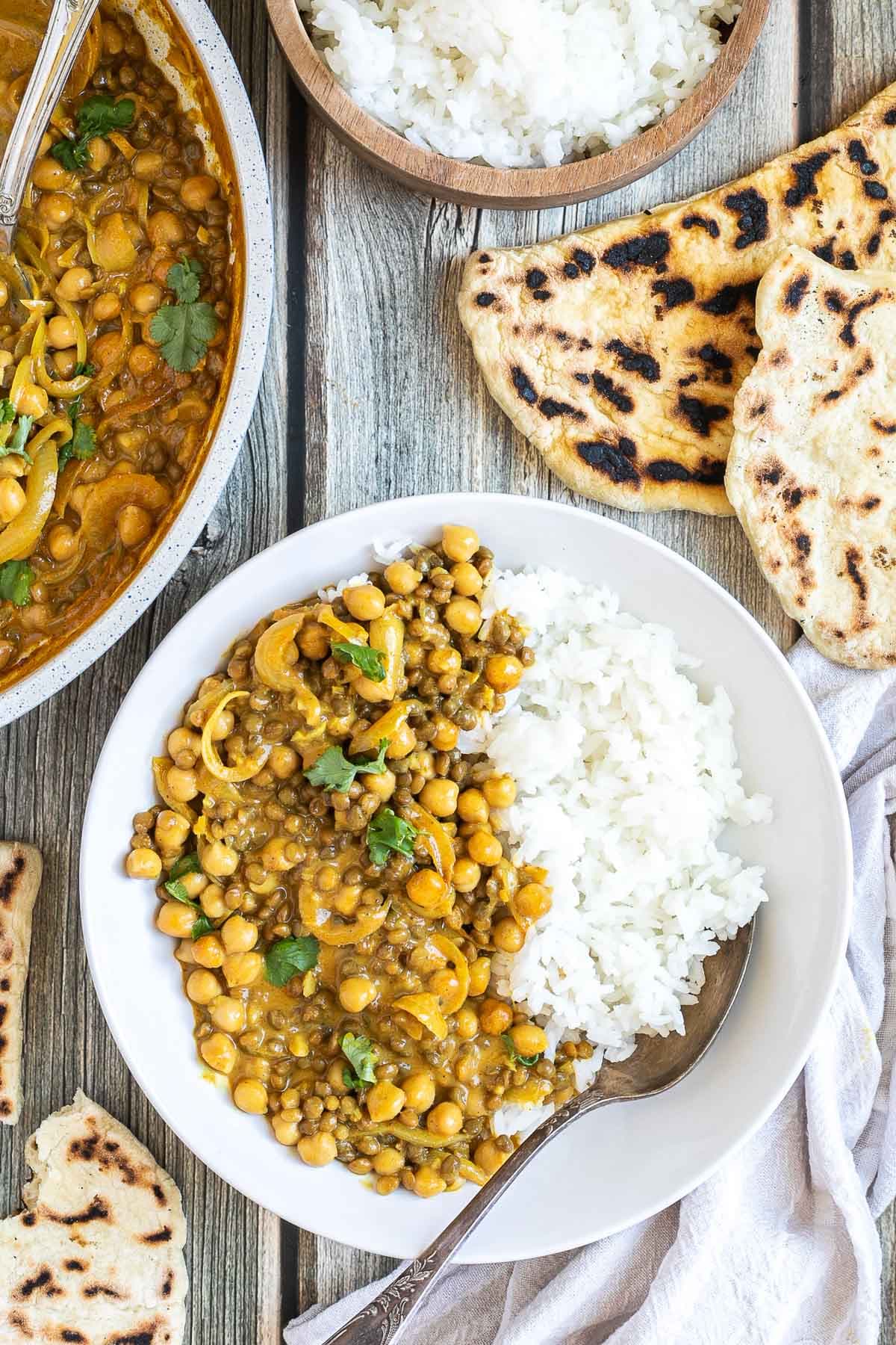 A white plate with white rice, chickpeas, and lentils, in a creamy light brown sauce sprinkled freshly chopped cilantro. A spoon is placed inside. Naan bread and the remaining curry in the pan are next to it.