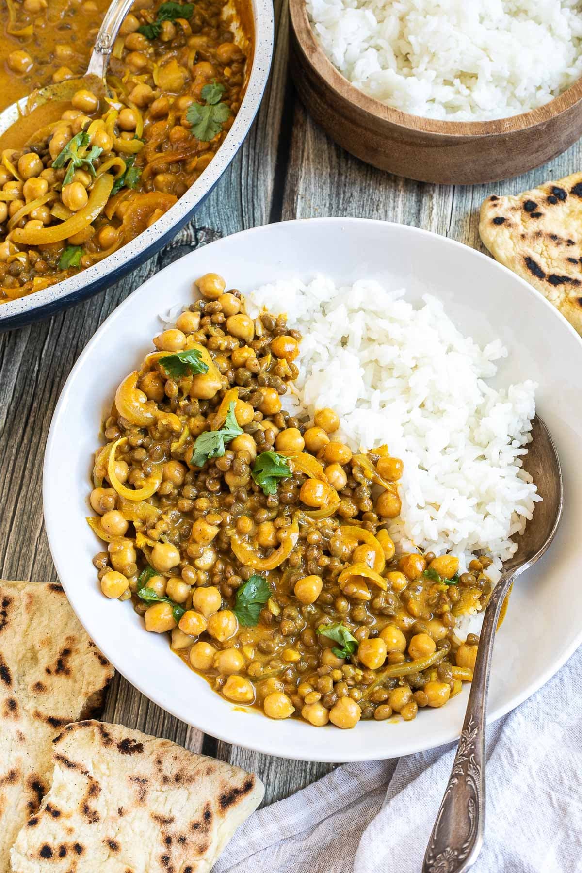 A white plate with white rice, chickpeas, and lentils, in a creamy light brown sauce sprinkled freshly chopped cilantro. A spoon is placed inside. Naan bread, a bowl of rice, and the remaining curry in the pan are next to it.