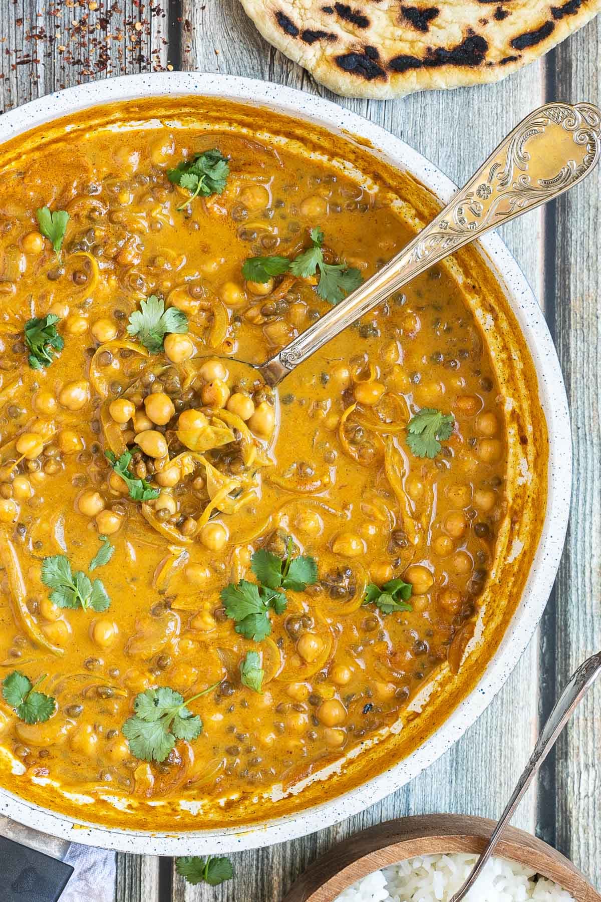 A white frying pan with chickpeas and lentils in a creamy light brown sauce sprinkled freshly chopped cilantro. A spoon is placed inside. Naan bread and a bowl of rice are next to it.