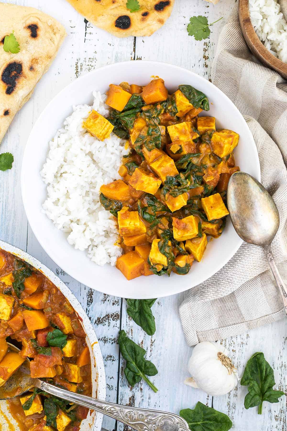 A white plate with white rice and tofu cubes, spinach leaves and orange sweet potato cubes in a thick orange curry sauce. Flatbread pieces are around it and the remaining curry in a white frying pan.