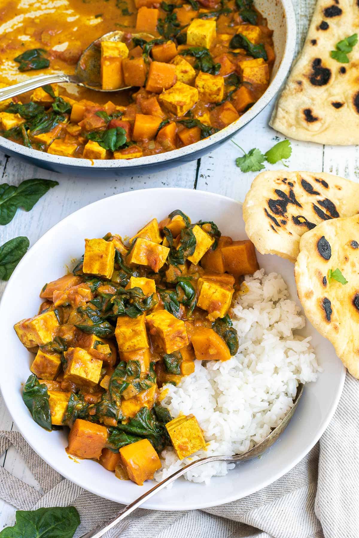 A white plate with white rice and tofu cubes, spinach leaves and orange sweet potato cubes in a thick orange curry sauce. Flatbread pieces are around it and the remaining curry in a white frying pan.