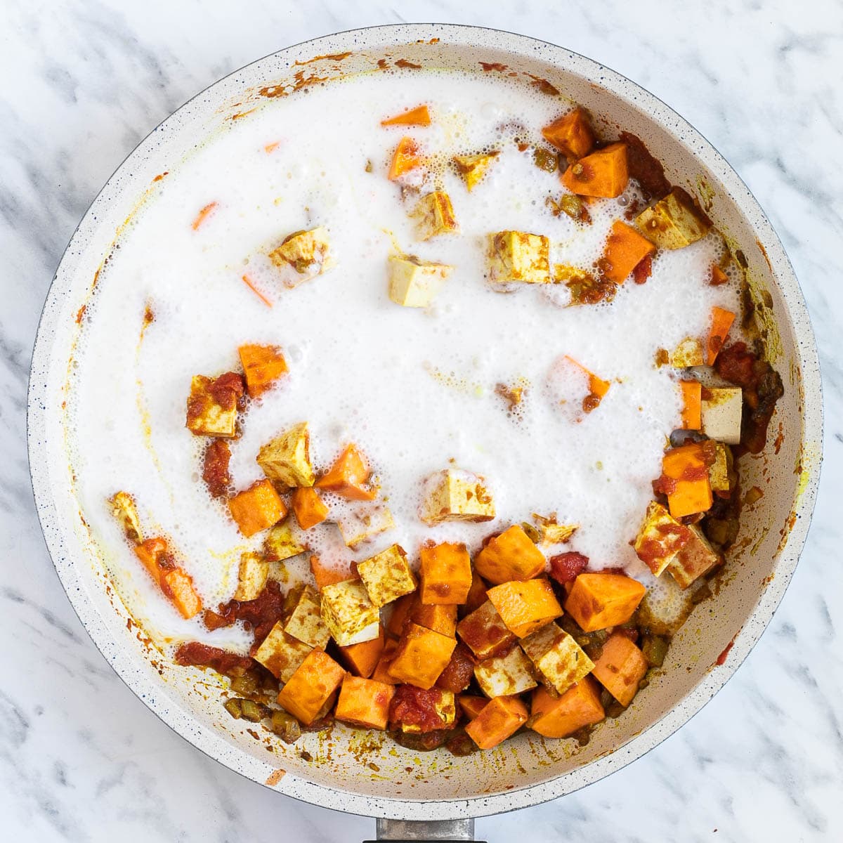 A white frying pan with tomato sauce, chopped orange sweet potato and white tofu cubes topped with white thick sauce, coconut cream.