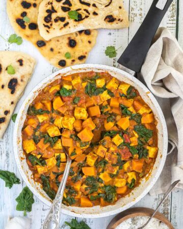 A white frying pan with tofu cubes, spinach leaves and orange sweet potato cubes in a thick orange curry sauce. Flatbread pieces are around it and a wooden bowl of white rice.