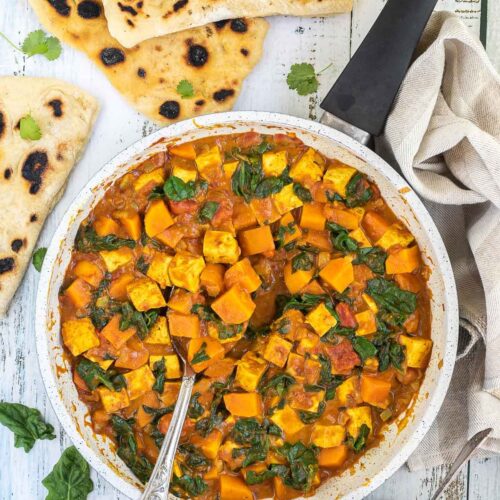 A white frying pan with tofu cubes, spinach leaves and orange sweet potato cubes in a thick orange curry sauce. Flatbread pieces are around it and a wooden bowl of white rice.
