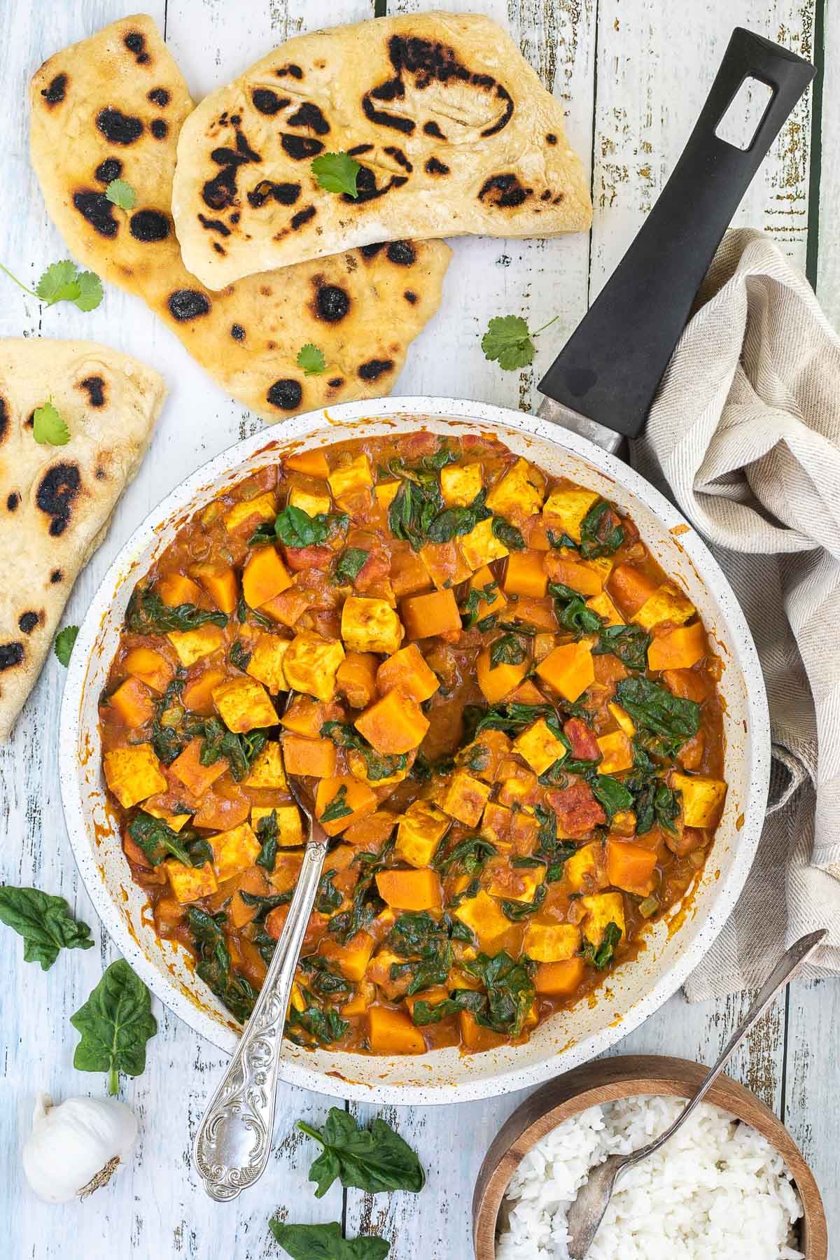 A white frying pan with tofu cubes, spinach leaves and orange sweet potato cubes in a thick orange curry sauce. Flatbread pieces are around it and a wooden bowl of white rice. 