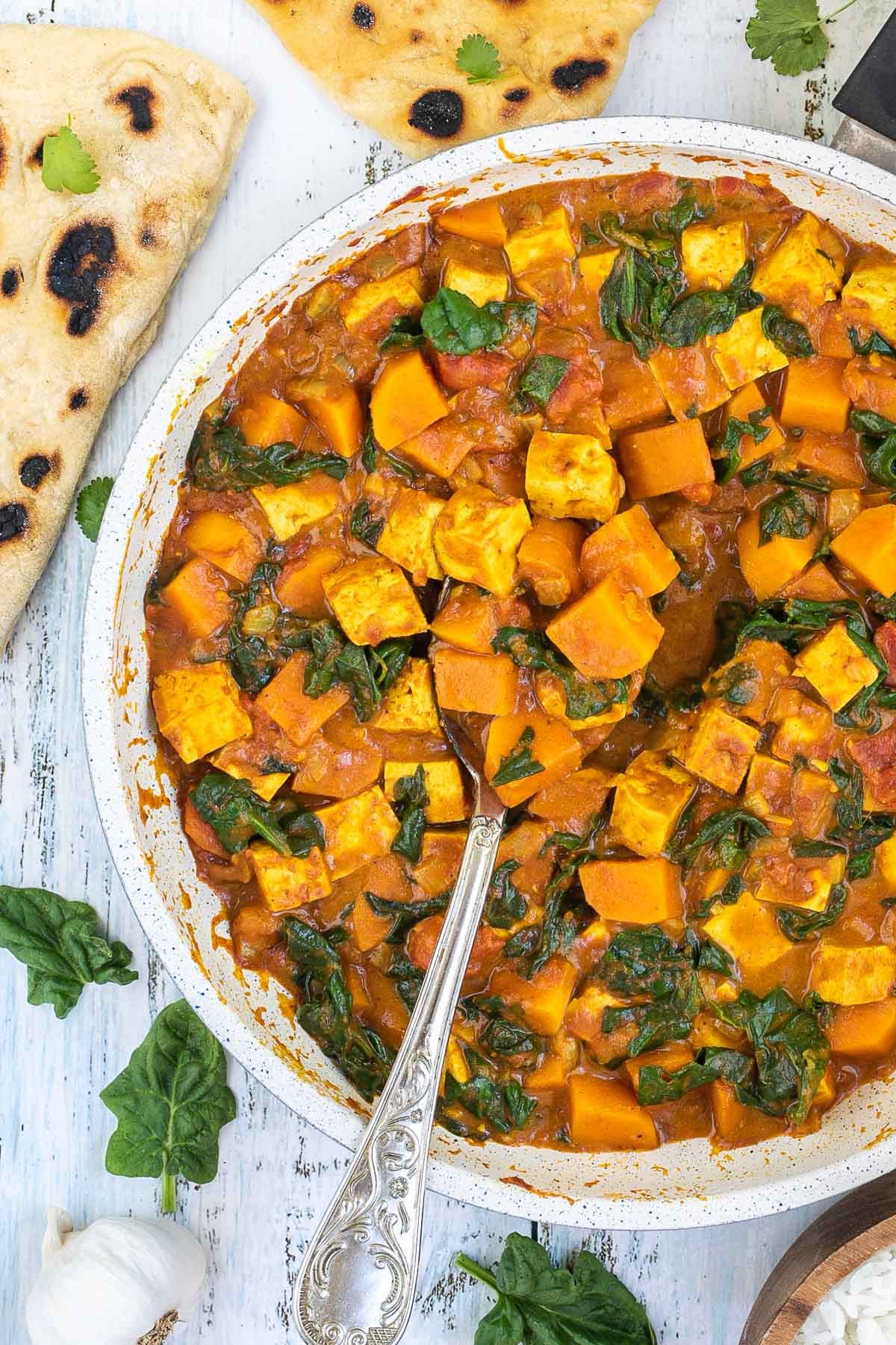 A white frying pan with tofu cubes, spinach leaves and orange sweet potato cubes in a thick orange curry sauce. Flatbread pieces are around it and a wooden bowl of white rice.