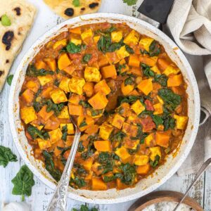 A white frying pan with tofu cubes, spinach leaves and orange sweet potato cubes in a thick orange curry sauce. Flatbread pieces are around it and a wooden bowl of white rice.