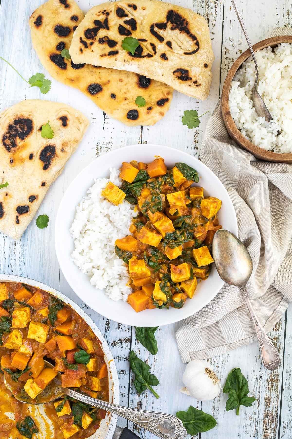 A white plate with white rice and tofu cubes, spinach leaves and orange sweet potato cubes in a thick orange curry sauce. Flatbread pieces are around it and the remaining curry in a white frying pan.