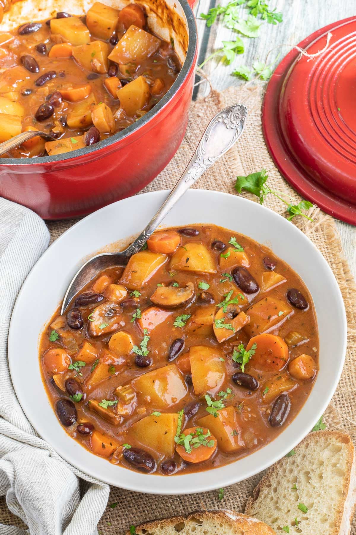 Vegetable stew in a white bowl with lots of chopped vegetables like potatoes, carrots, mushroom, celery and red kidney beans. Topped with freshly chopped green herbs. A spoon is placed in the middle.