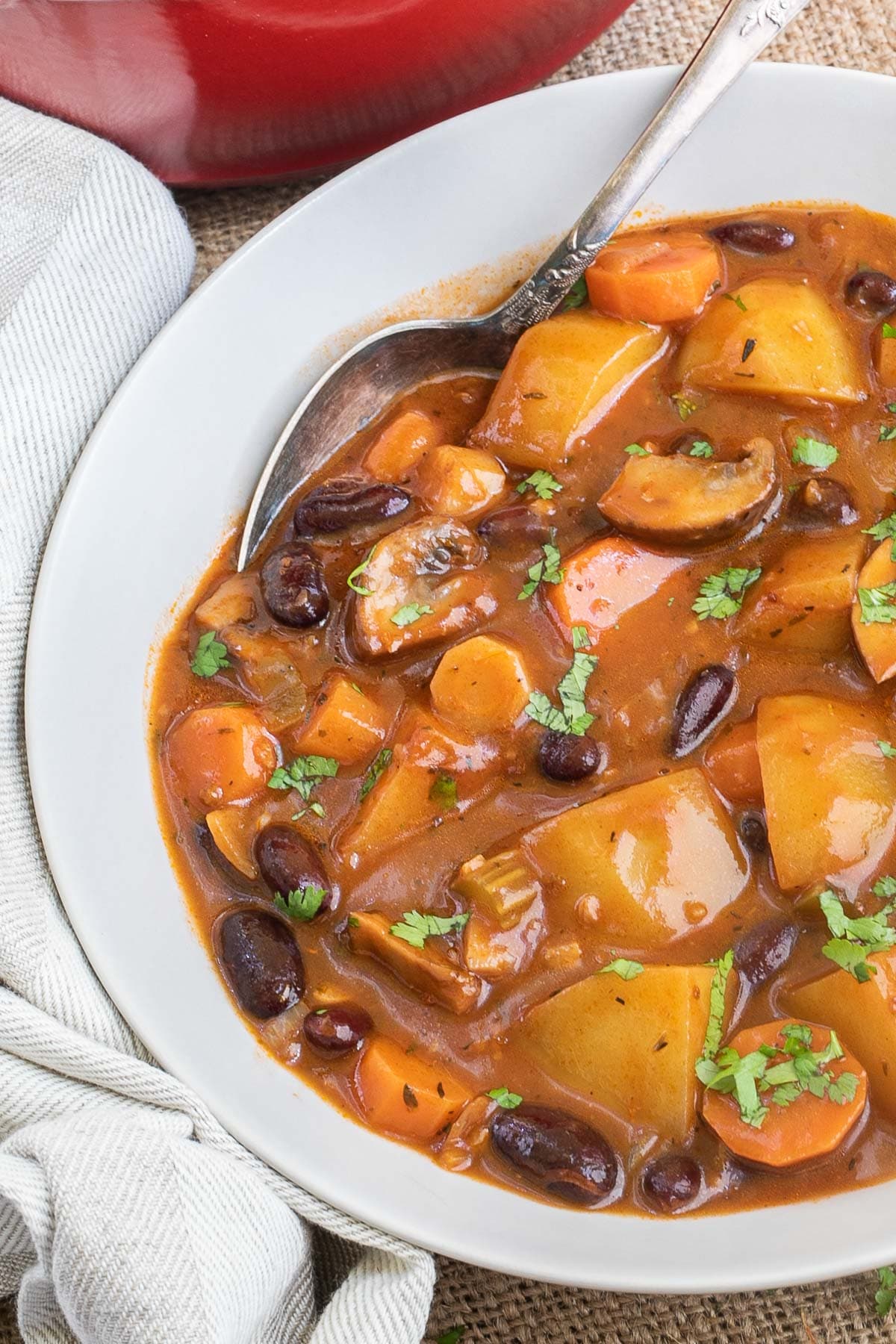 Vegetable stew in a white bowl with lots of chopped vegetables like potatoes, carrots, mushroom, celery and red kidney beans. Topped with freshly chopped green herbs. A spoon is placed in the middle.