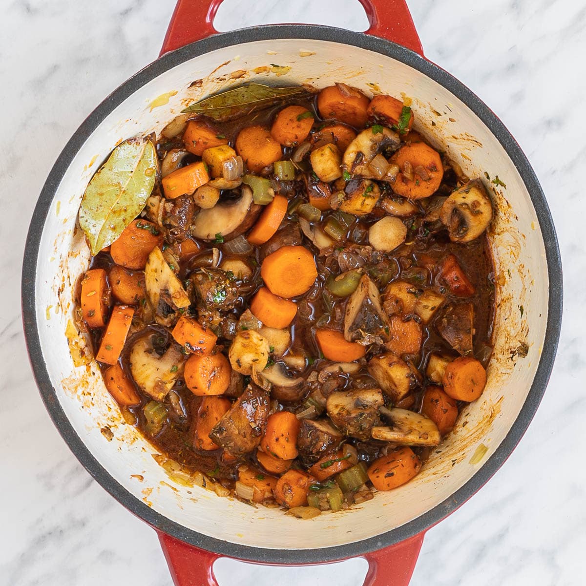 Red white enameled Dutch oven with lots of chopped veggies like carrots, mushrooms, celery and small heaps of green herbs and red spices