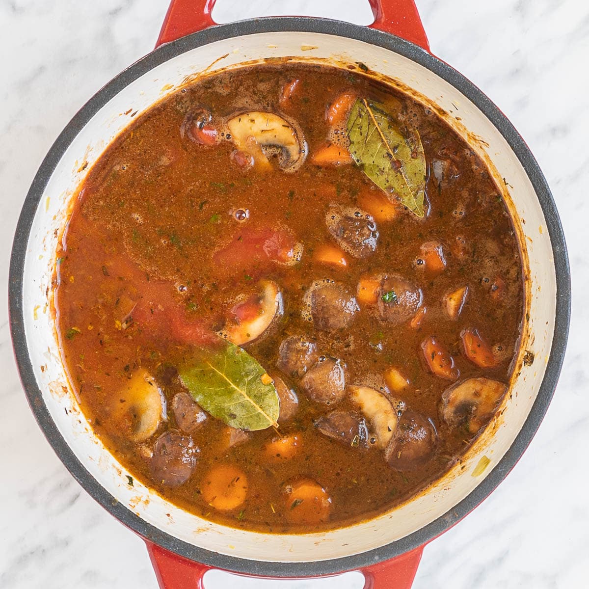 Red white enameled Dutch oven with a red thick sauce, lots of chopped veggies like carrots, mushrooms, celery.