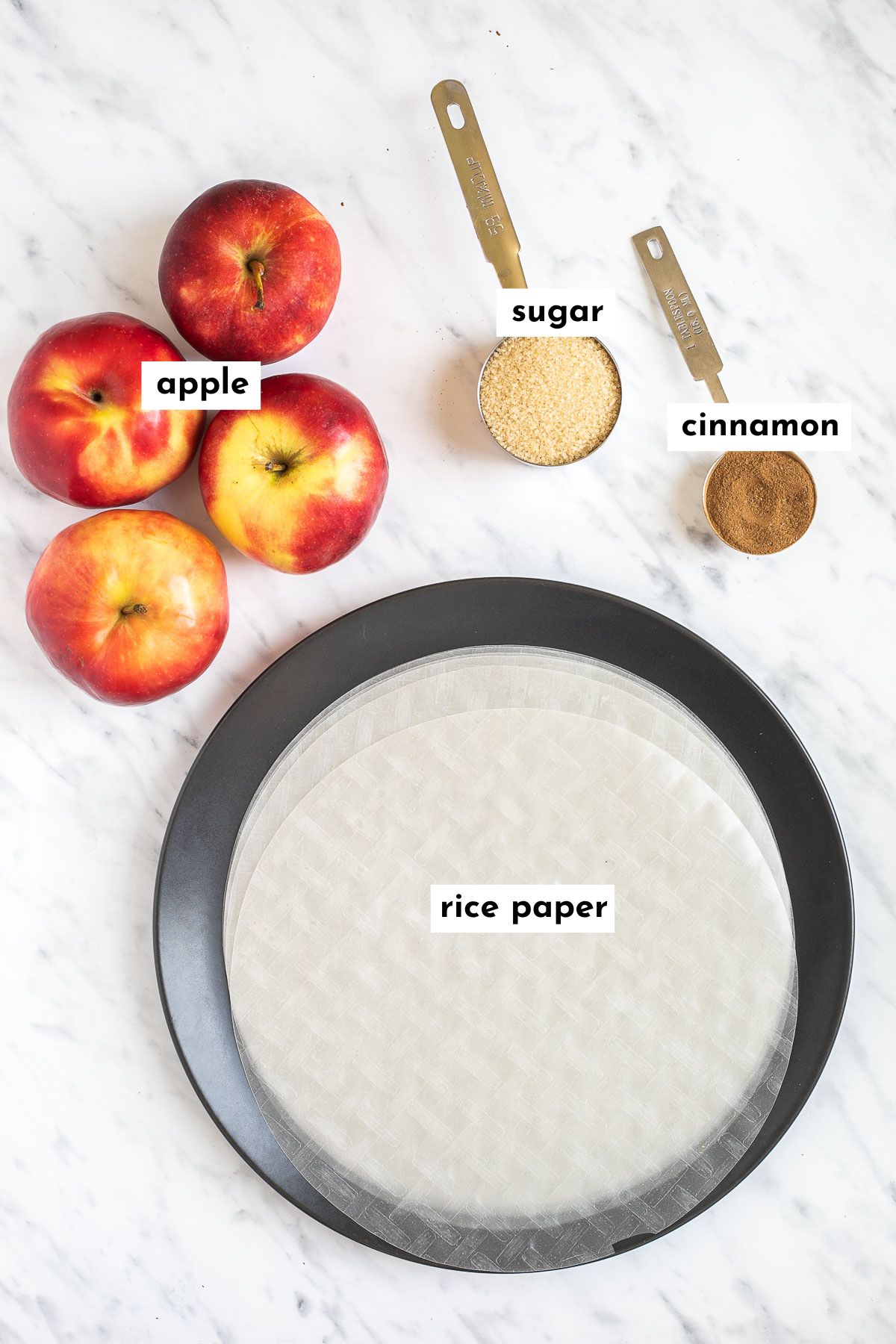 Ingredients of apple pie spring rolls placed on a marble surface. They are apples, rice paper sheets, a measuring spoon of sugar and a measuring spoon full of cinnamon.