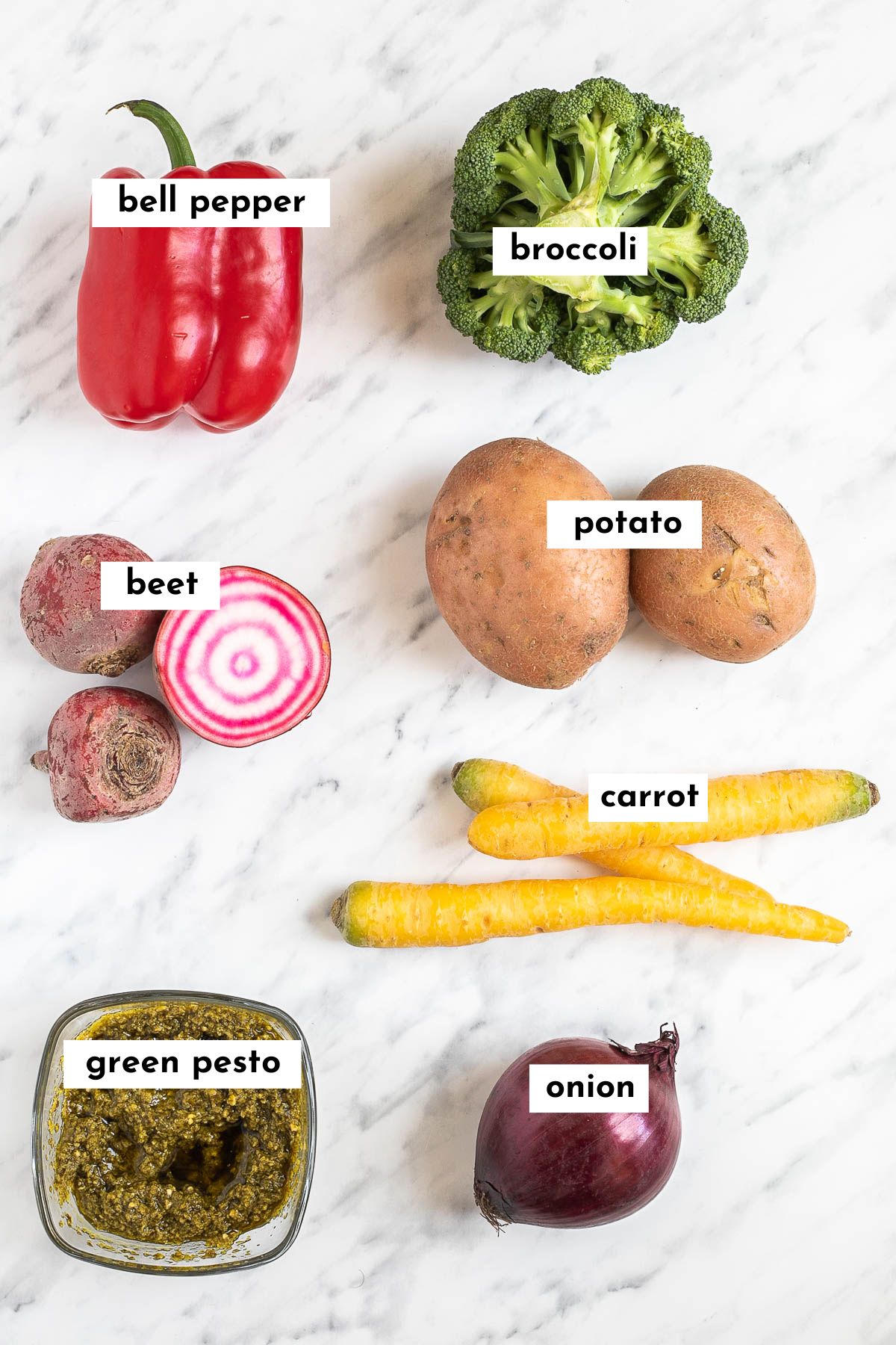 The ingredients of pesto vegetables on a marble countertop red bell pepper, broccoli, pink beet, potatoes, yellow carrot, red onion and green pesto sauce.