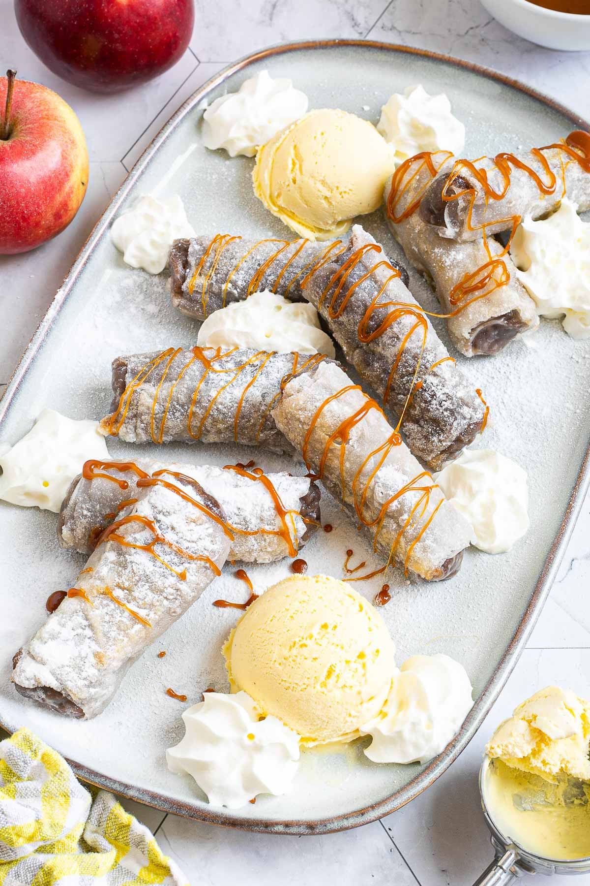Rice paper rolls with brown filling placed on a light blue plate. Dusted with powdered sugar. Served with scoops of vanilla ice cream, caramel drizzle, and heaps of whipped cream.