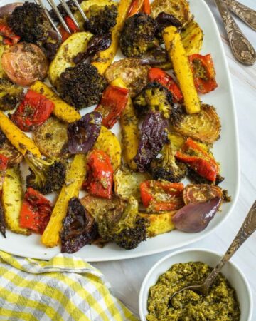 Large white serving plate with vibrant vegetables like broccoli, yellow carrots, red pepper, red onion, pink beet and potato slightly charred around the edges covered in a bit of pesto sauce. A fork is taking on stick.