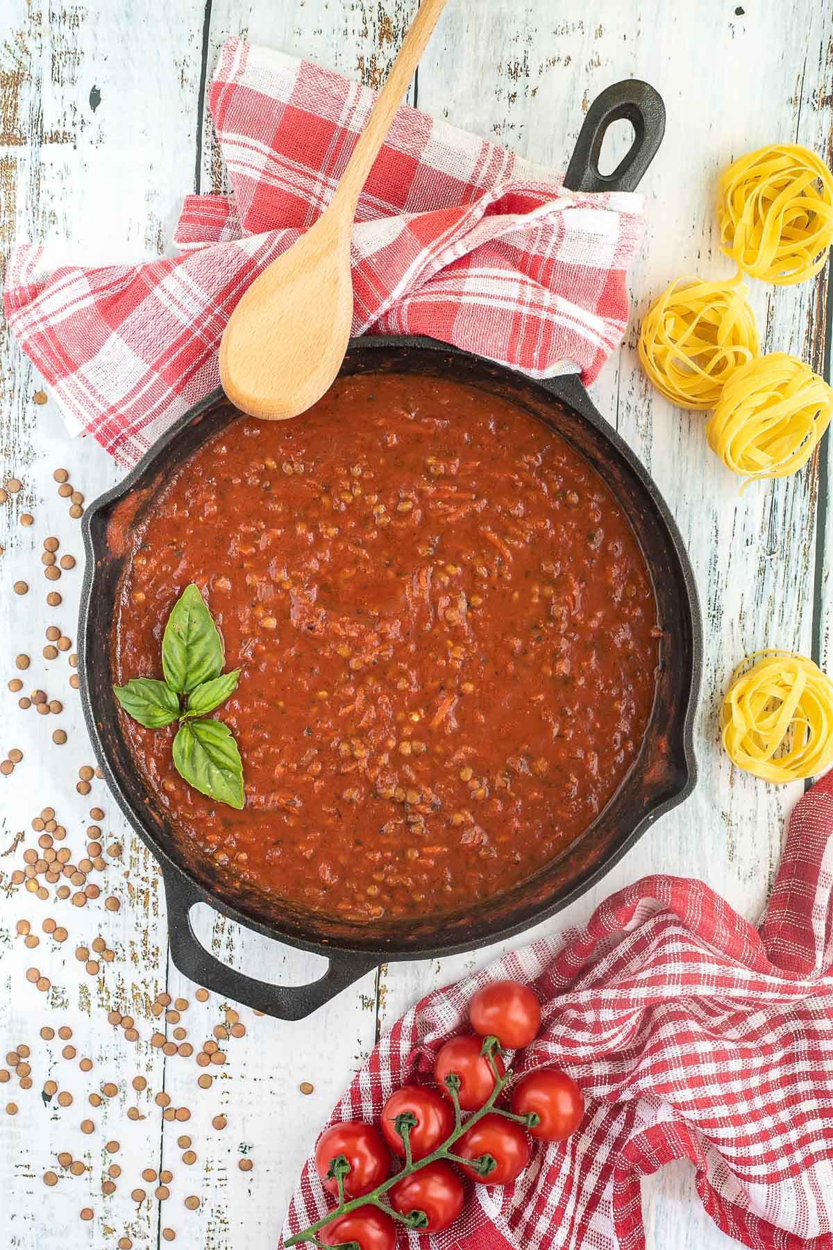 A cast iron skillet with brown lentils in bolognese sauce. Dry lentils, dry pasta and some cherry tomatoes are scattered around the skillet. A wooden spatula is placed on the side.