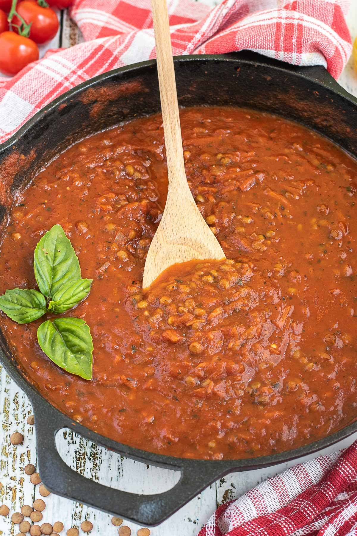 A cast iron skillet with brown lentils in bolognese sauce. Dry lentils, dry pasta and some cherry tomatoes are scattered around the skillet. A wooden spatula is placed in the middle of the sauce.