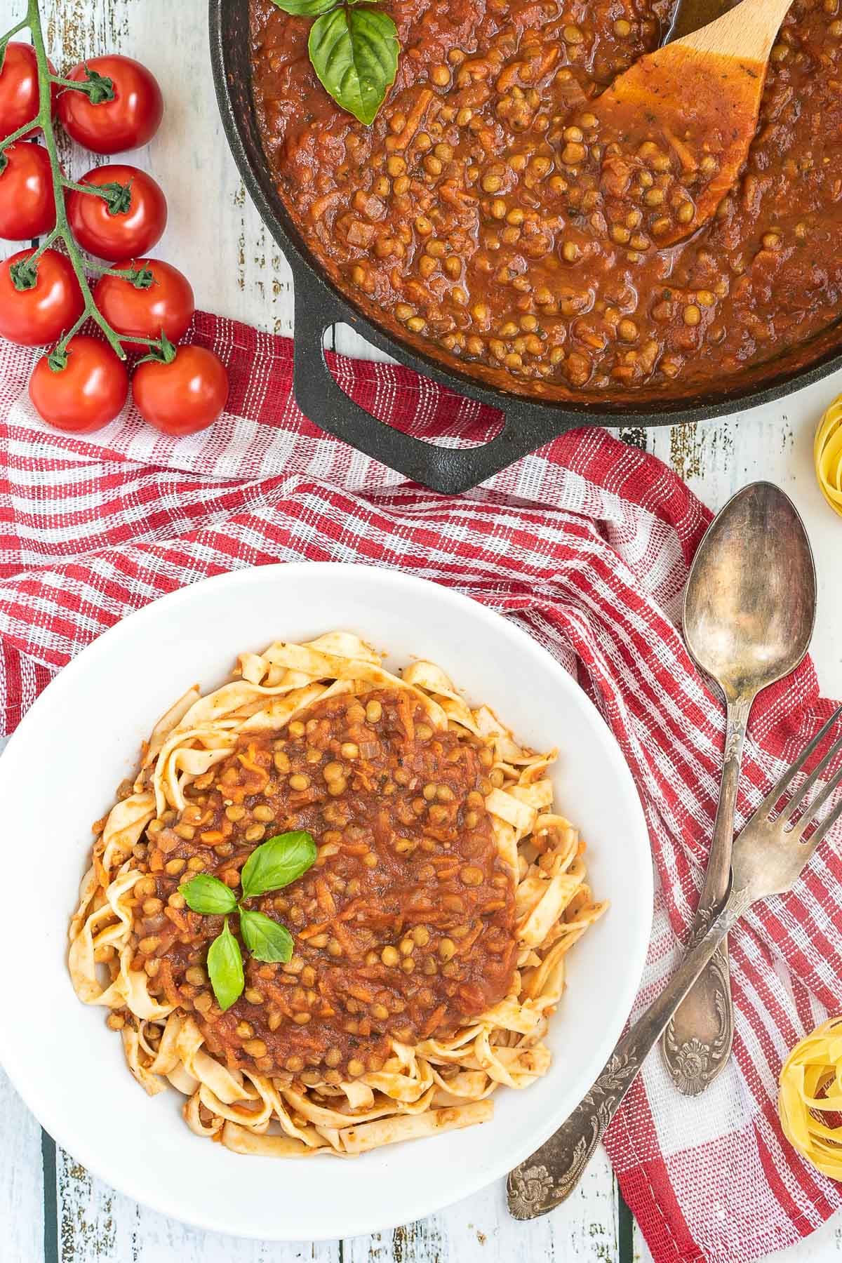 A white bowl of tagliatelle pasta topped with lentils in bolognese sauce. Leftover bolognese in the cast iron skillet is right next to it.