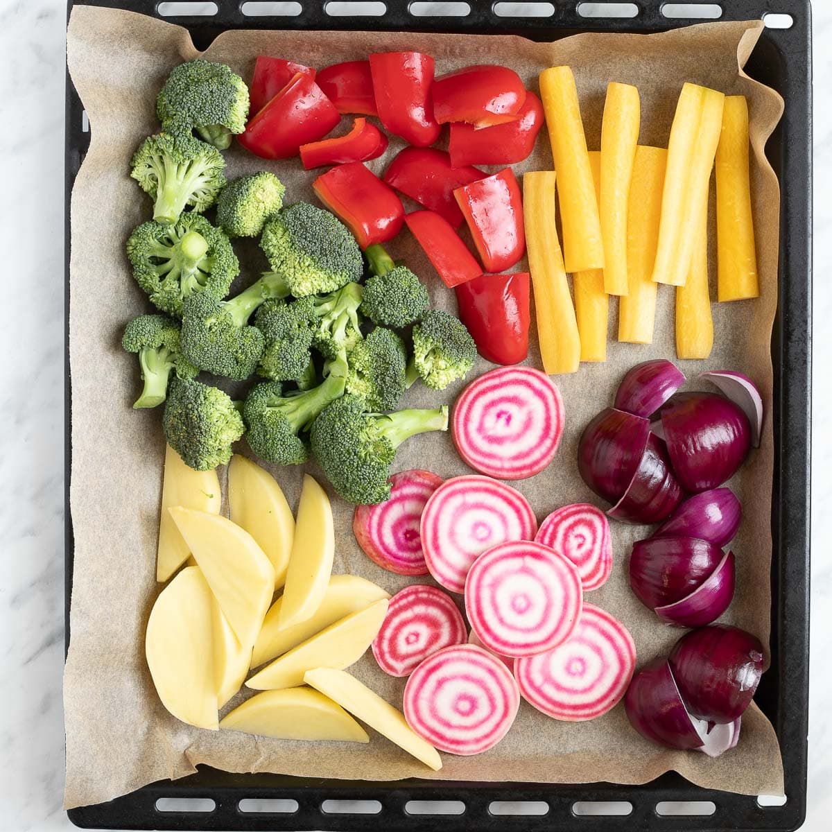 A large black sheet pan covered with parchment pepper with raw veggies like red bell pepper, broccoli, pink beet, potatoes, yellow carrot, red onion and green pesto sauce.