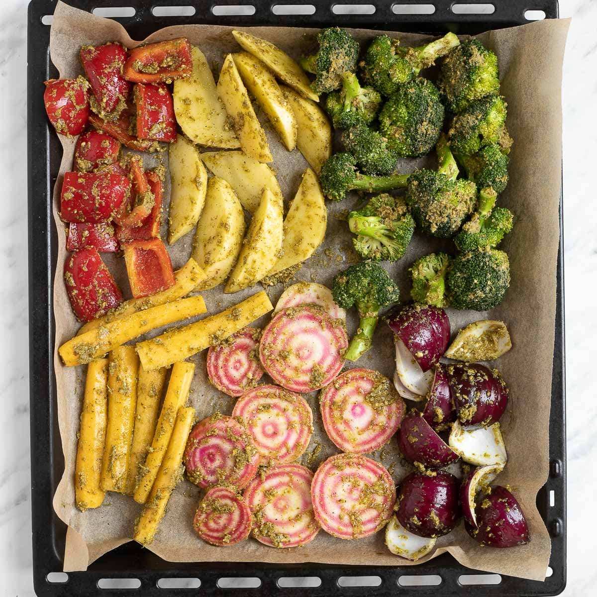 A large black sheet pan covered with parchment pepper with raw veggies like red bell pepper, broccoli, pink beet, potatoes, yellow carrot, red onion and green pesto sauce covered in green pesto.