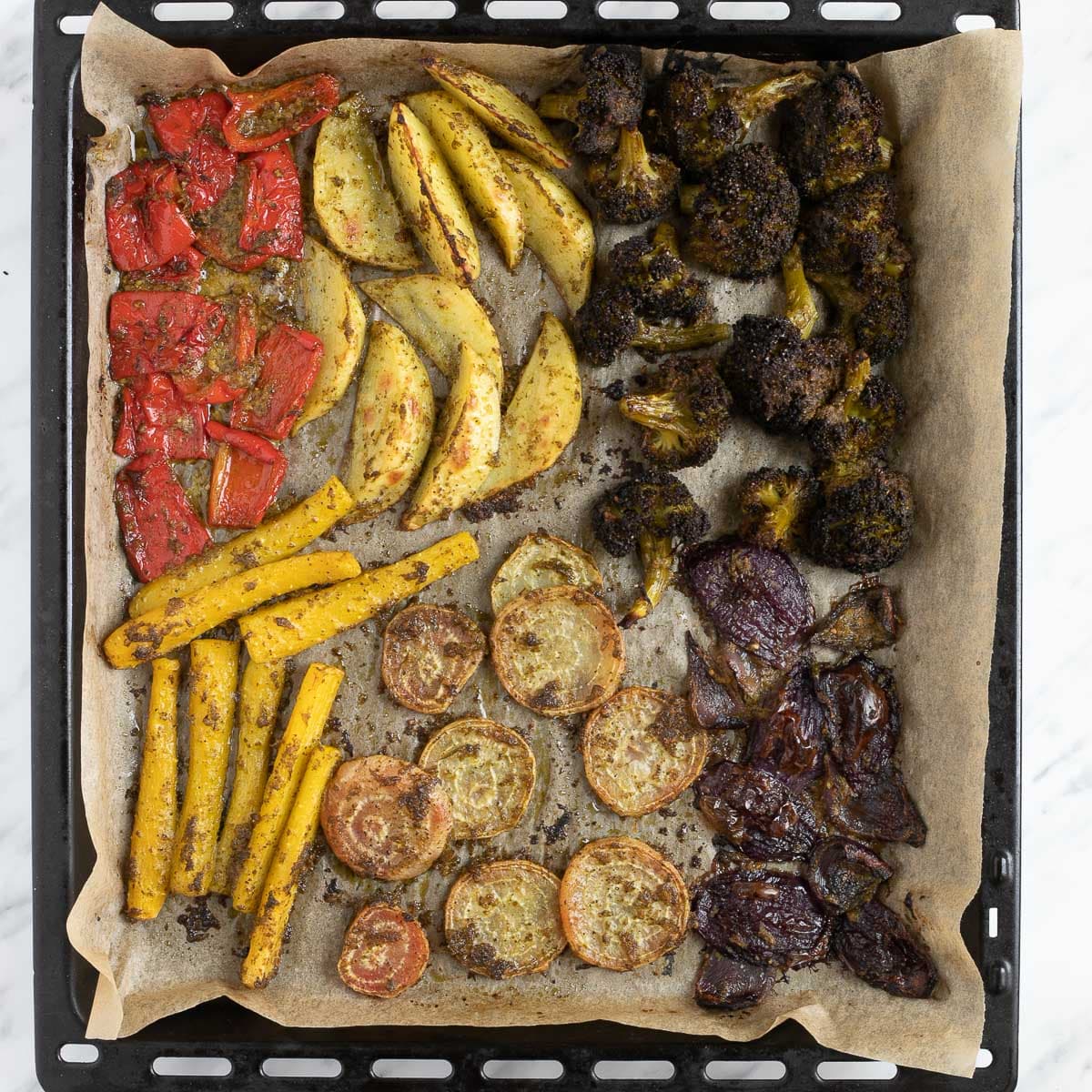 A large black sheet pan covered with parchment pepper with roasted veggies like red bell pepper, broccoli, pink beet, potatoes, yellow carrot, red onion and green pesto sauce covered in green pesto.