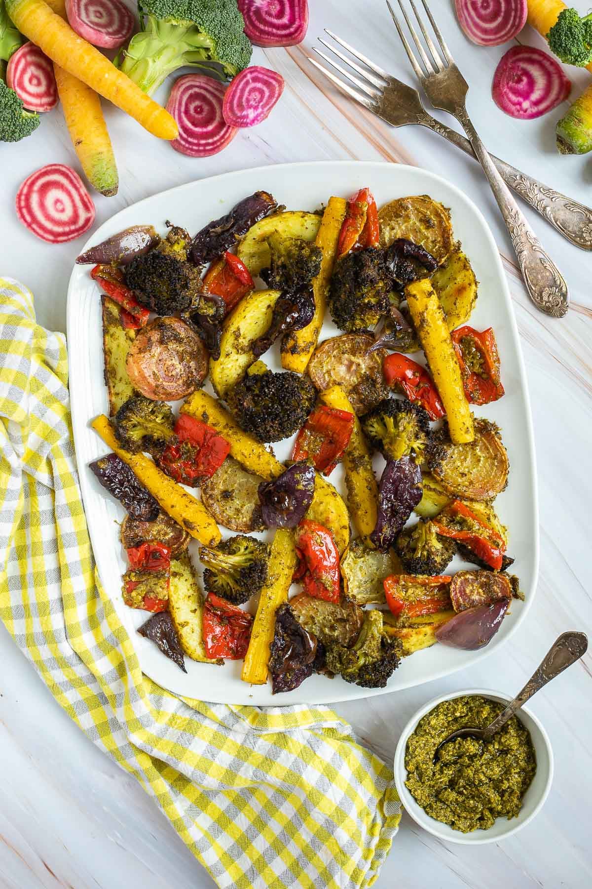 Large white serving plate with vibrant vegetables like broccoli, yellow carrots, red pepper, red onion, pink beet and potato slightly charred around the edges covered in a bit of pesto sauce. Extra pesto and raw veggies are next to it.