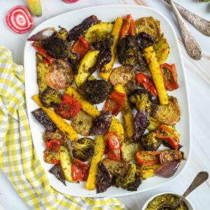 Large white serving plate with vibrant vegetables like broccoli, yellow carrots, red pepper, red onion, pink beet and potato slightly charred around the edges covered in a bit of pesto sauce. Extra pesto and raw veggies are next to it.