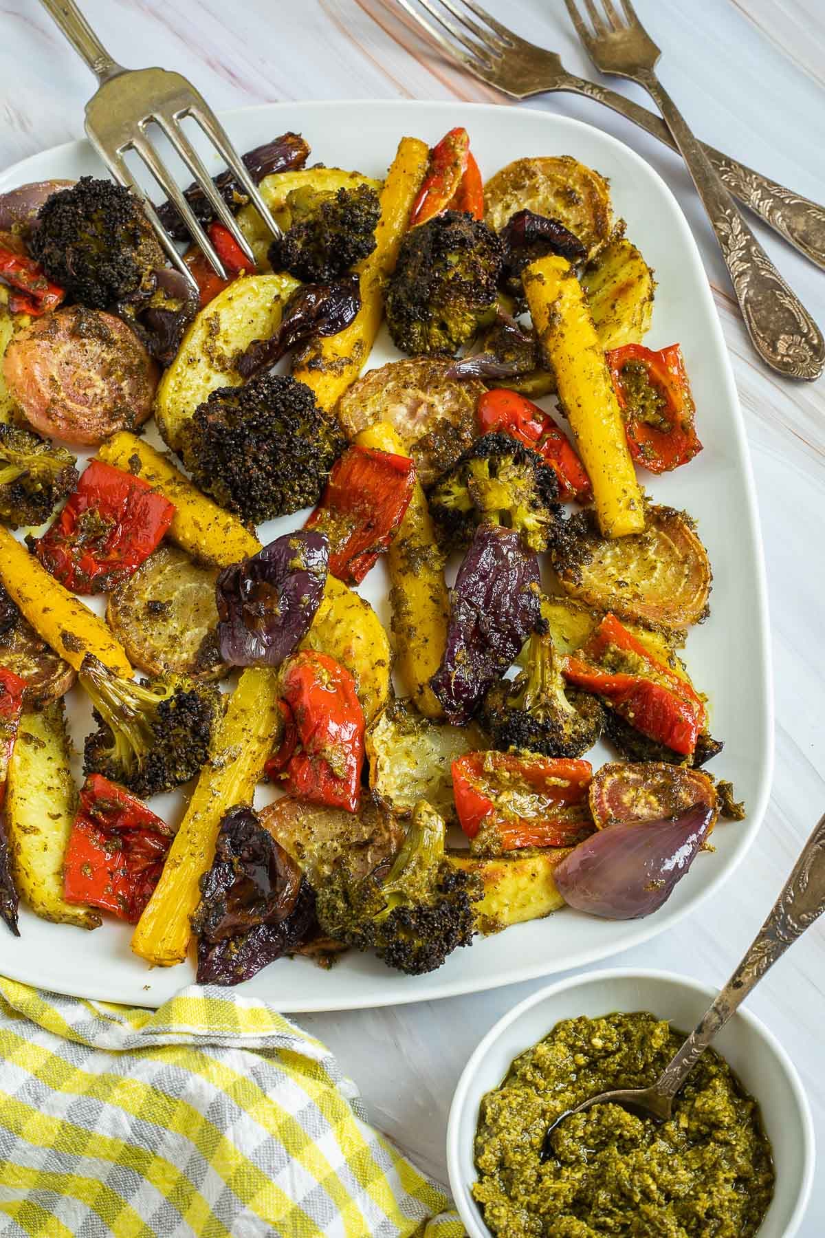 Large white serving plate with vibrant vegetables like broccoli, yellow carrots, red pepper, red onion, pink beet and potato slightly charred around the edges covered in a bit of pesto sauce. A fork is taking on stick.