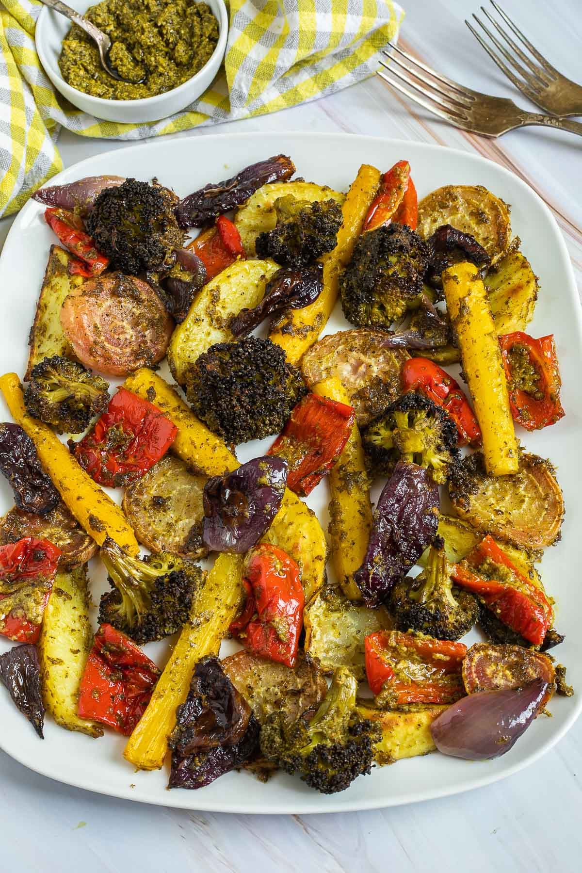 Large white serving plate with vibrant vegetables like broccoli, yellow carrots, red pepper, red onion, pink beet and potato slightly charred around the edges covered in a bit of pesto sauce. Extra pesto and raw veggies are next to it.