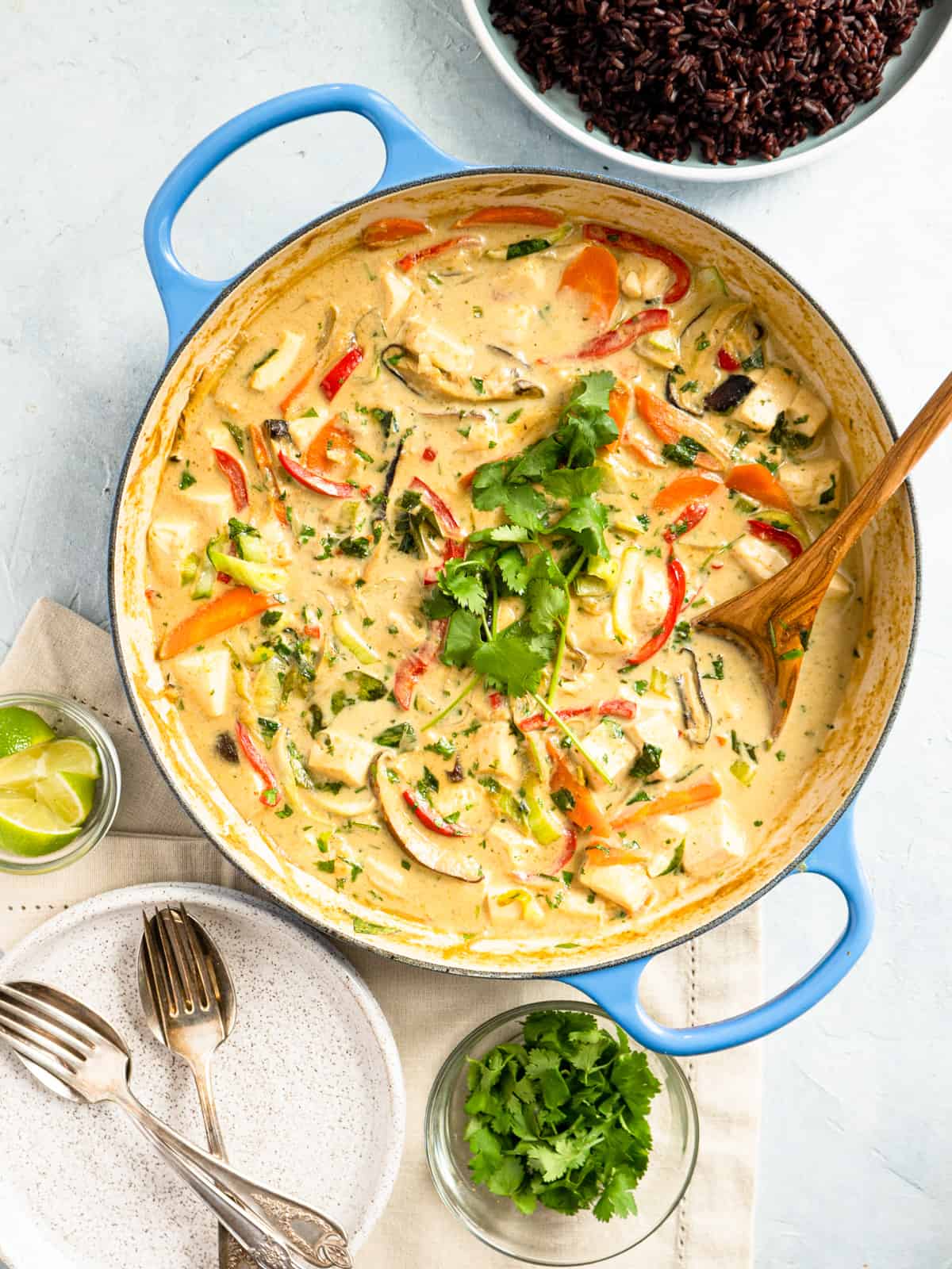 one pot of prepared green curry with plates, fresh cilantro, lime wedges, and black rice on the side. 