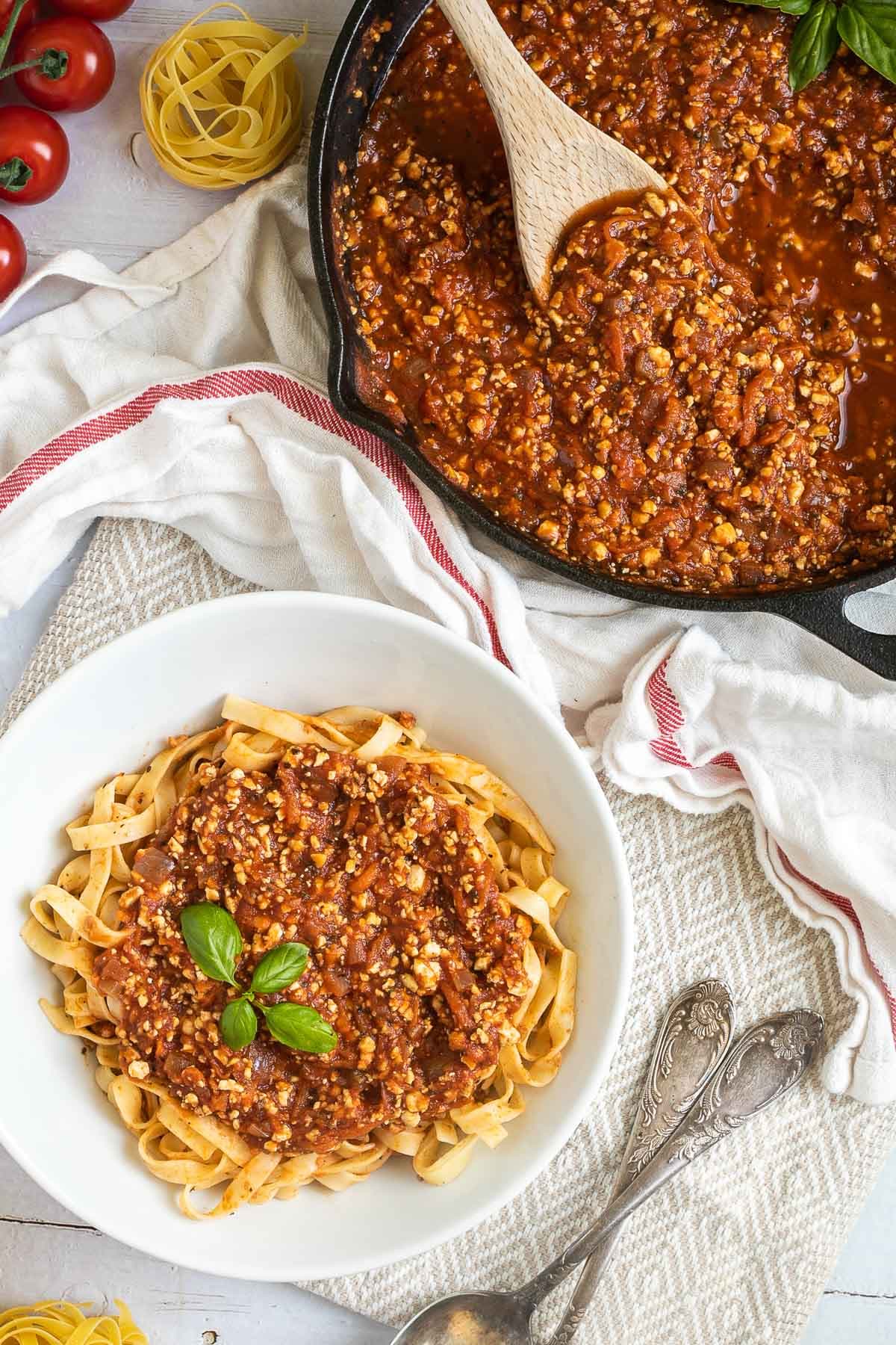 A white bowl of tagliatelle pasta topped with tofu mince in bolognese sauce. Leftover bolognese in the cast iron skillet is right next to it.