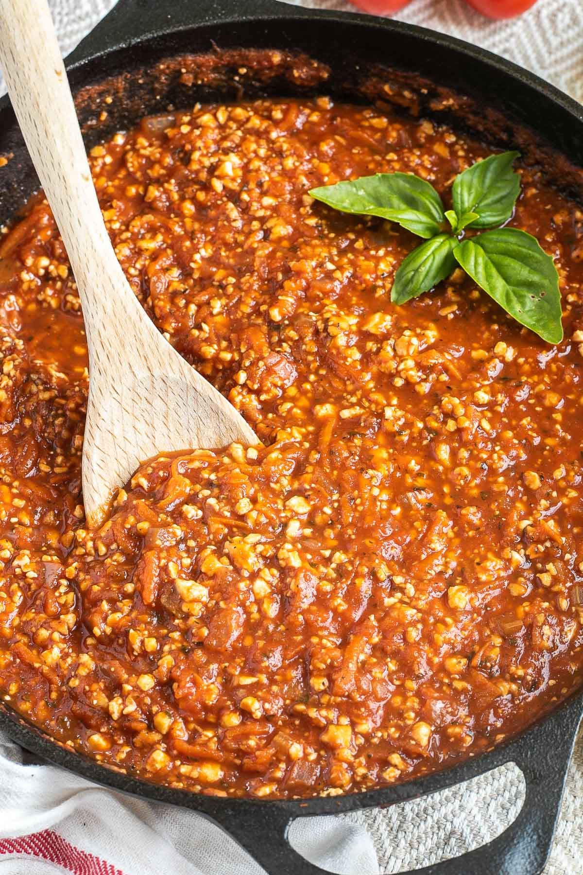 A cast iron skillet with tofu mince in bolognese sauce. A wooden spoon is placed in the middle.