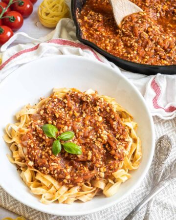A white bowl of tagliatelle pasta topped with tofu mince in bolognese sauce. Leftover bolognese in the cast iron skillet is right next to it.