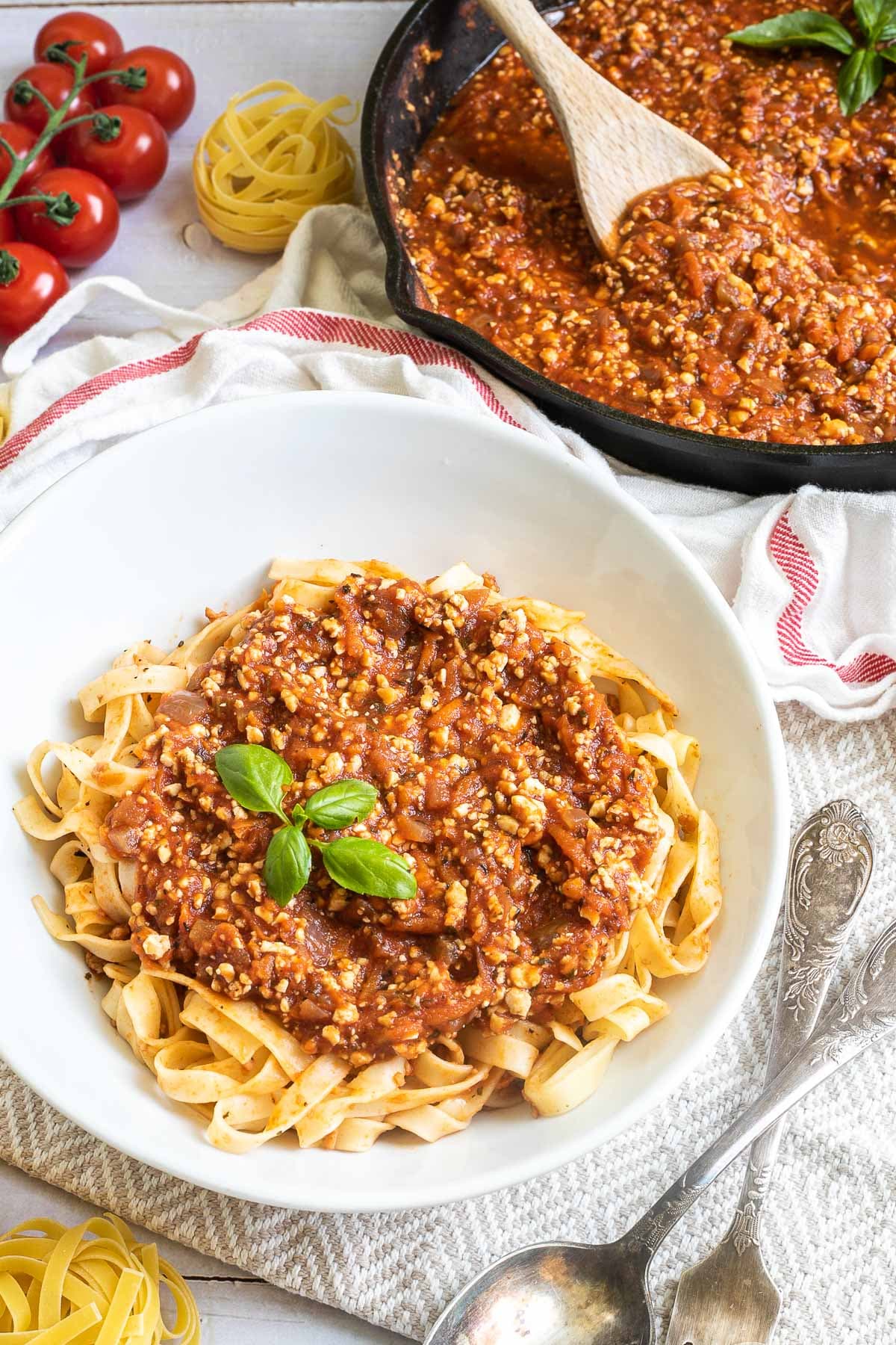 A white bowl of tagliatelle pasta topped with tofu mince in bolognese sauce. Leftover bolognese in the cast iron skillet is right next to it.