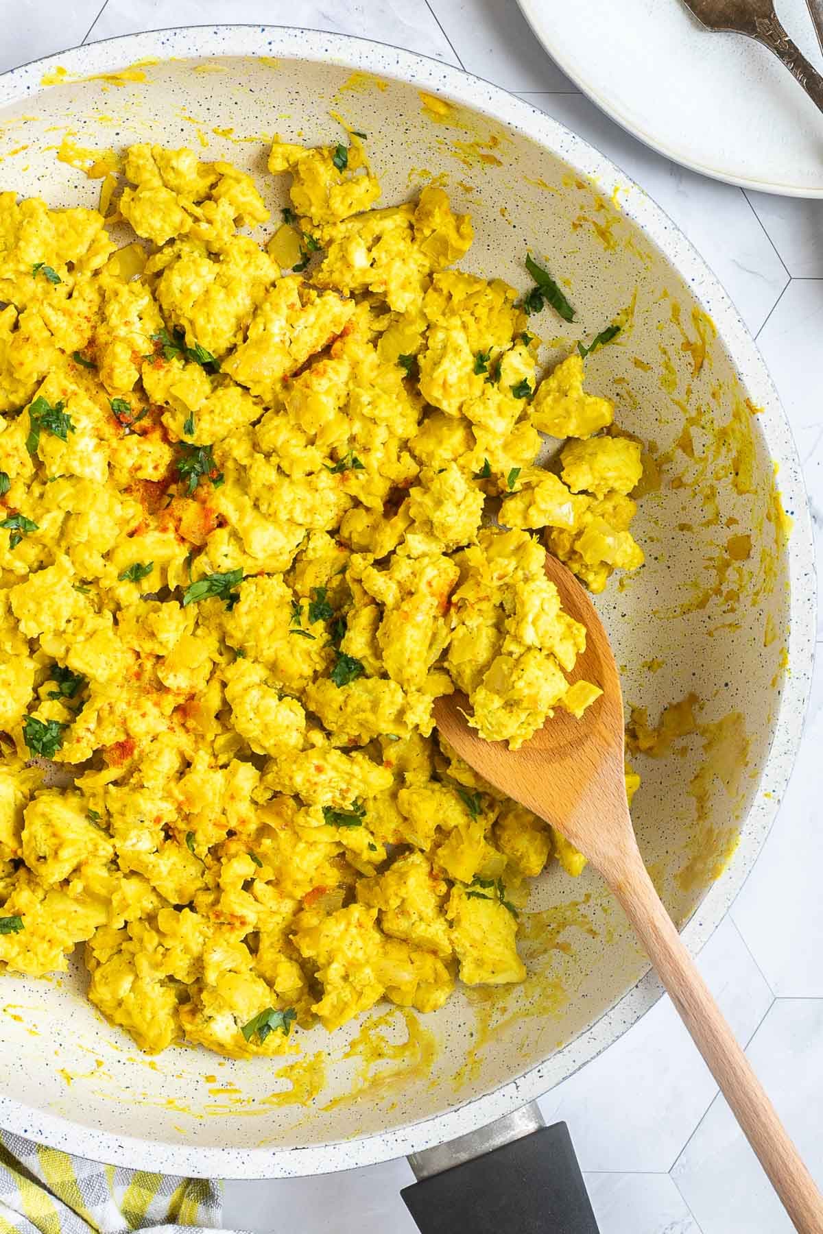 Yellow tofu crumbles in a white frying pan sprinkled with fresh herbs and red pepper flakes. A wooden spatula is placed inside.