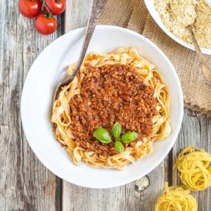 A white bowl of tagliatelle pasta topped with meat-like crumbles in bolognese sauce. Parmesan cheese in a small white bowl right next to it.