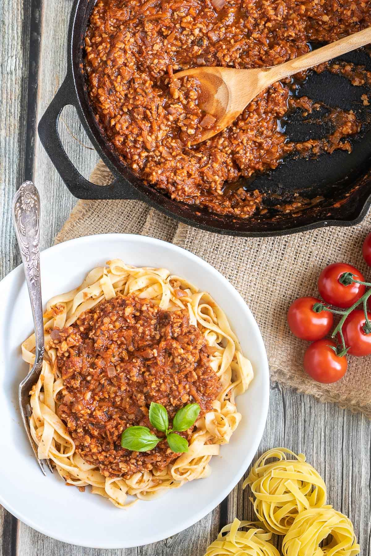 A white bowl of tagliatelle pasta topped with meat-like crumbles in bolognese sauce. Leftover bolognese in the cast iron skillet is right next to it.