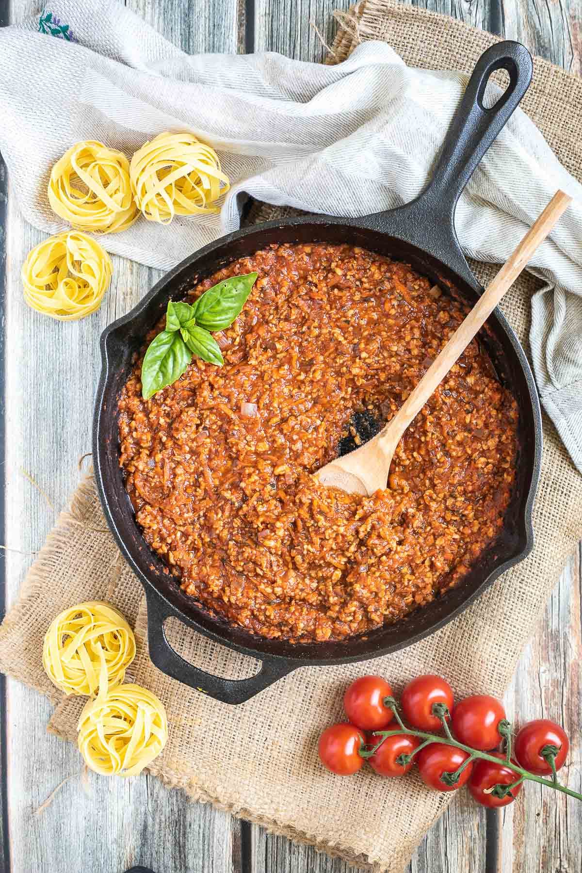 A cast iron skillet with meat-like crumbles in bolognese sauce. A wooden spatula is placed in the middle. Dry pasta and cherry tomatoes are around it.