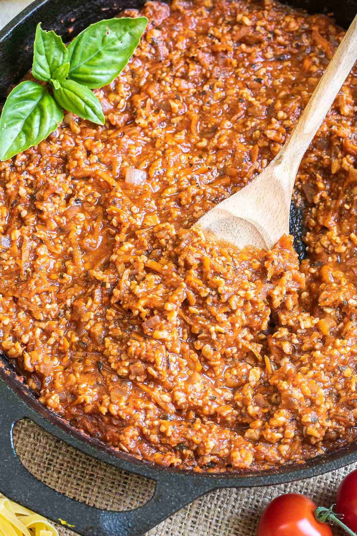 A cast iron skillet with meat-like crumbles in bolognese sauce. A wooden spatula is placed in the middle.