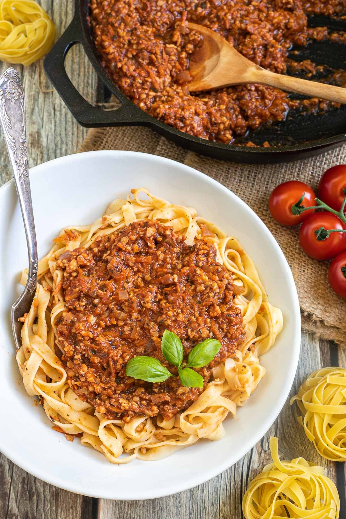 A white bowl of tagliatelle pasta topped with meat-like crumbles in bolognese sauce. Leftover bolognese in the cast iron skillet is right next to it.