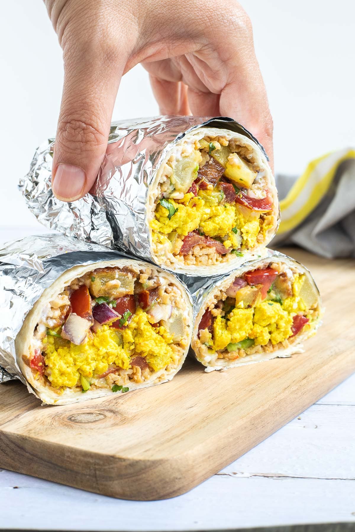 3 half tortillas wrapped in tin foil filled with sausage crumbles, diced potatoes, tofu scrambles, and avocado tomato salsa. A hand is taking the one from the top.