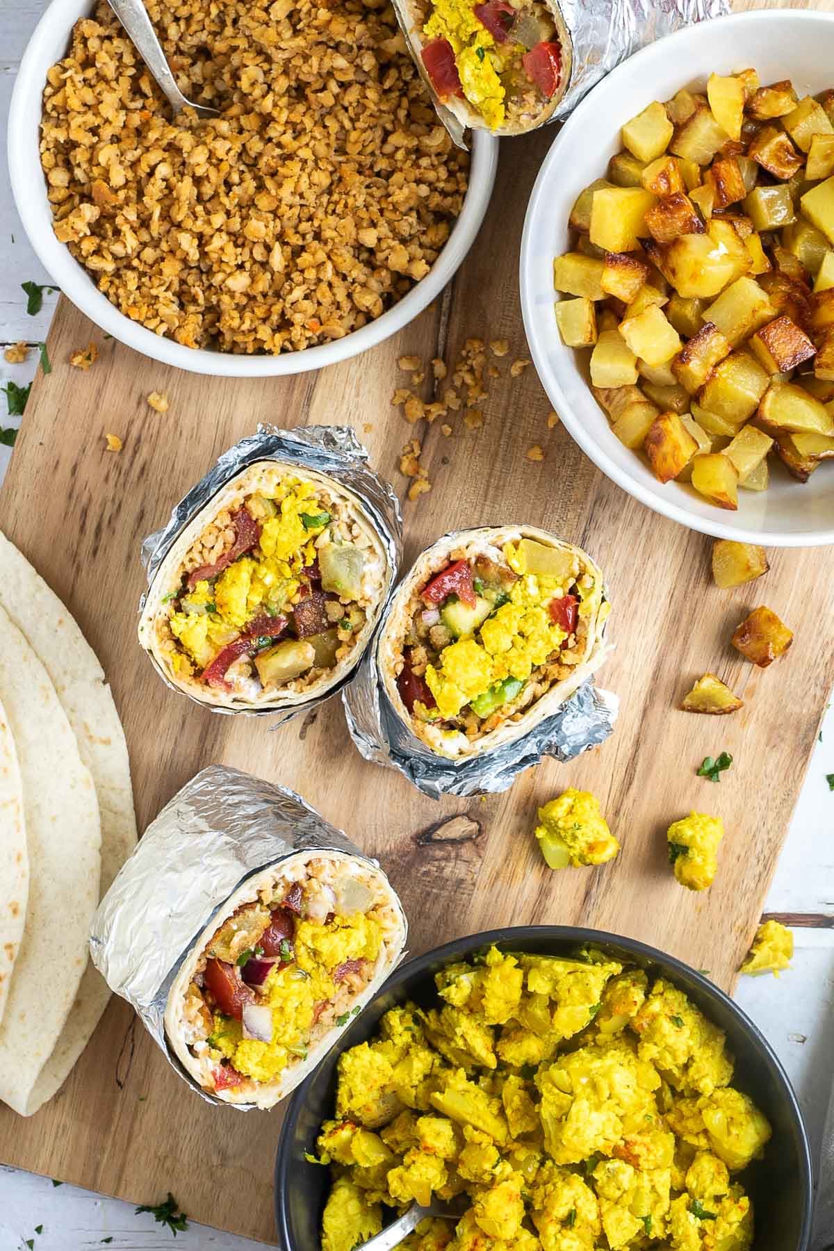 3 half tortillas wrapped in tin foil filled with sausage crumbles, diced potatoes, tofu scrambles, and avocado tomato salsa. The remaining fillings are placed in several bowls on a wooden platter.