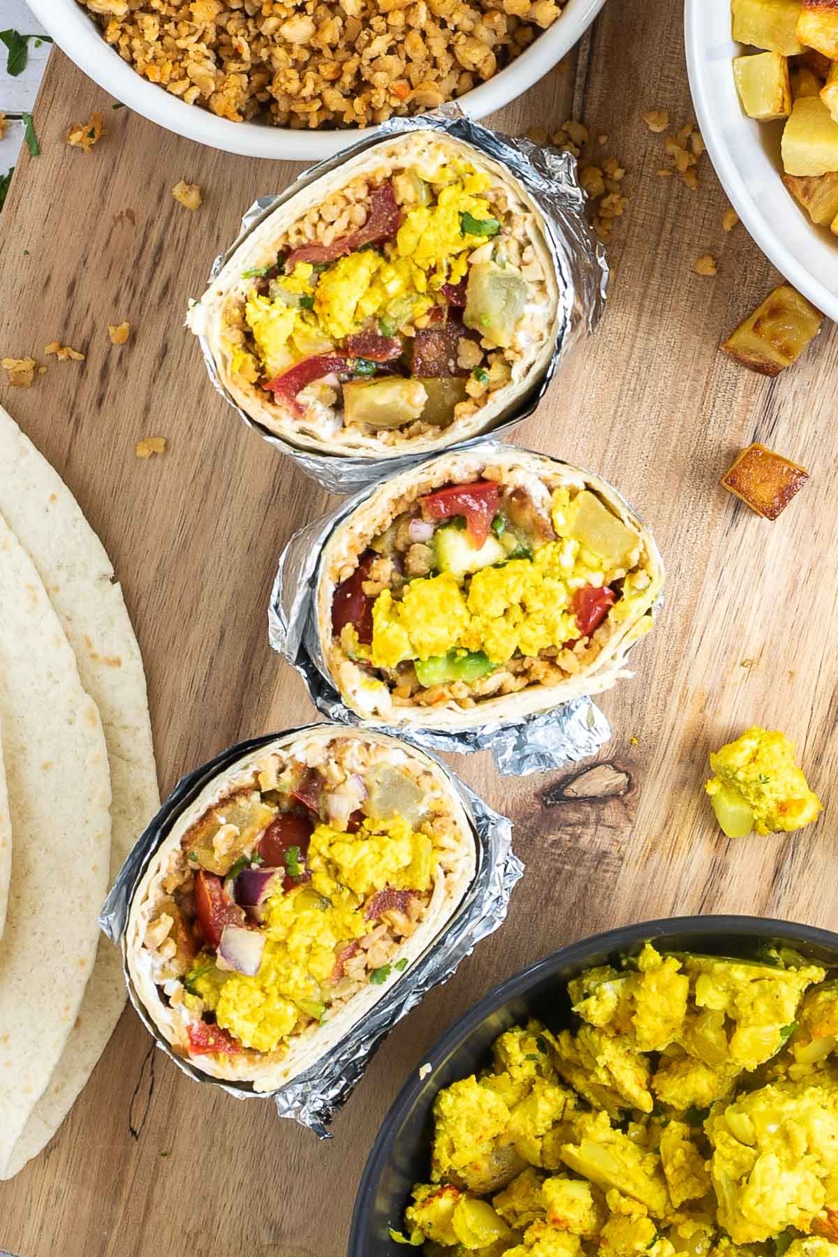 3 half tortillas wrapped in tin foil filled with sausage crumbles, diced potatoes, tofu scrambles, and avocado tomato salsa. The remaining fillings are placed in several bowls on a wooden platter.